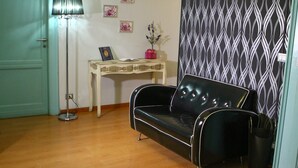 Una habitación con un sofá de cuero negro, suelo de madera, una lámpara de pie, una pequeña mesa con un florero y un libro, y una pared con un diseño decorativo.