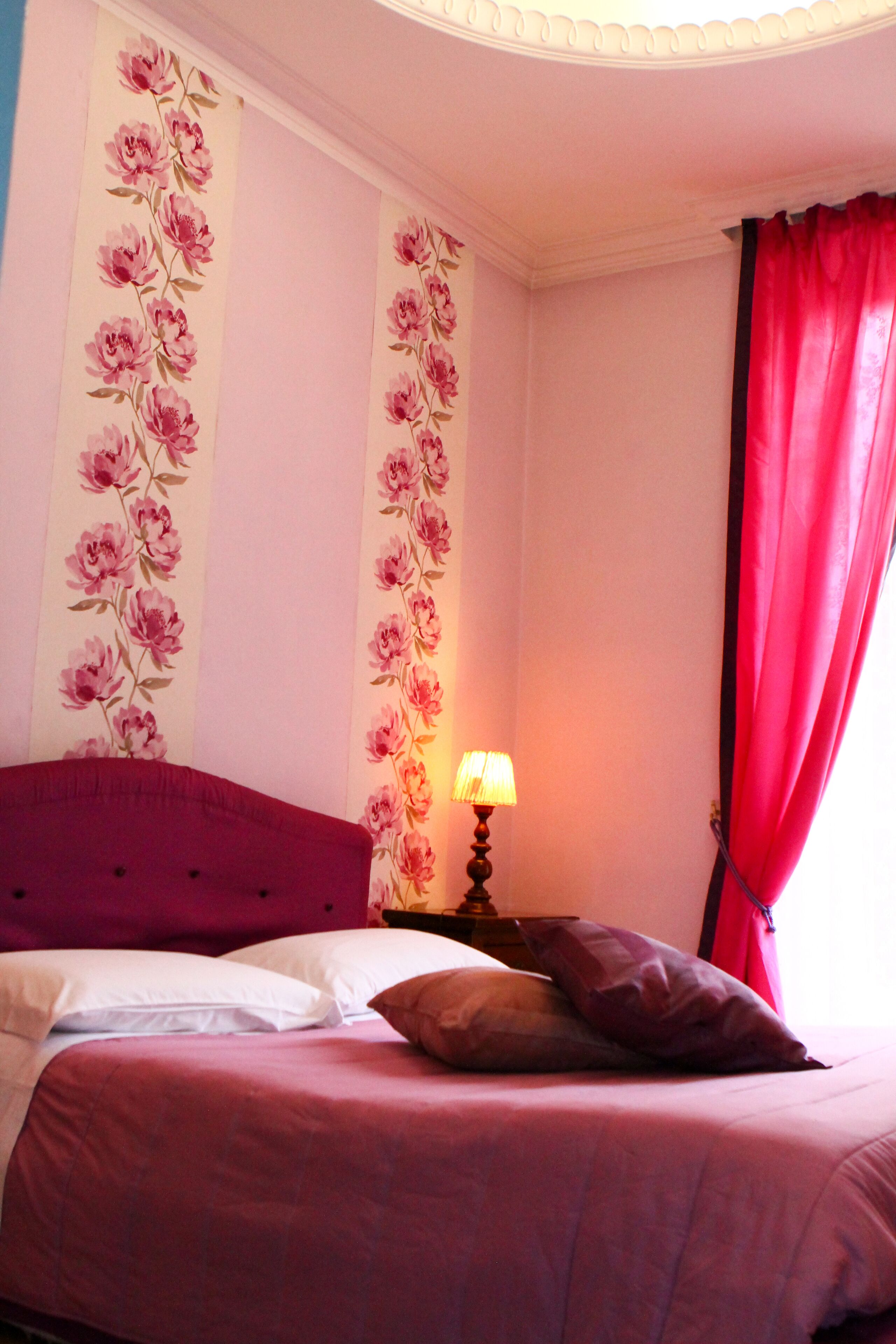 A bedroom with a bed, pillows, a lamp, and floral wallpaper.