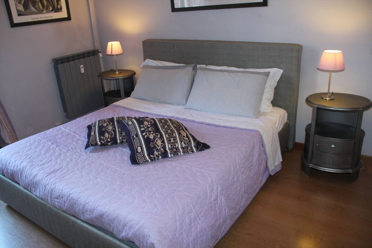 A neatly made bed with a patterned pillow, two bedside tables with lamps, and a radiator on the wall.
