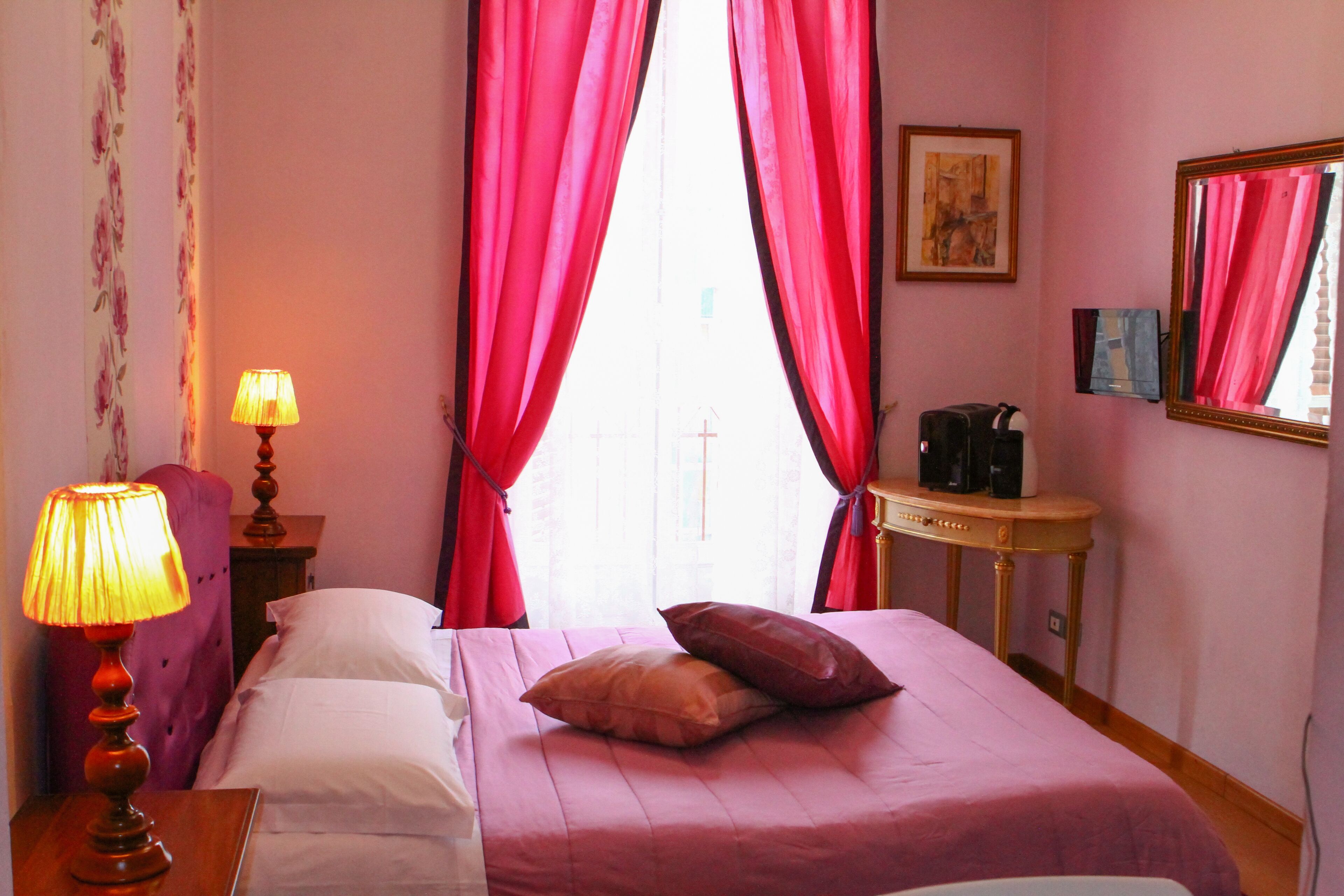 A hotel room with a bed, a nightstand with a lamp, a table with a coffee maker, and red curtains.