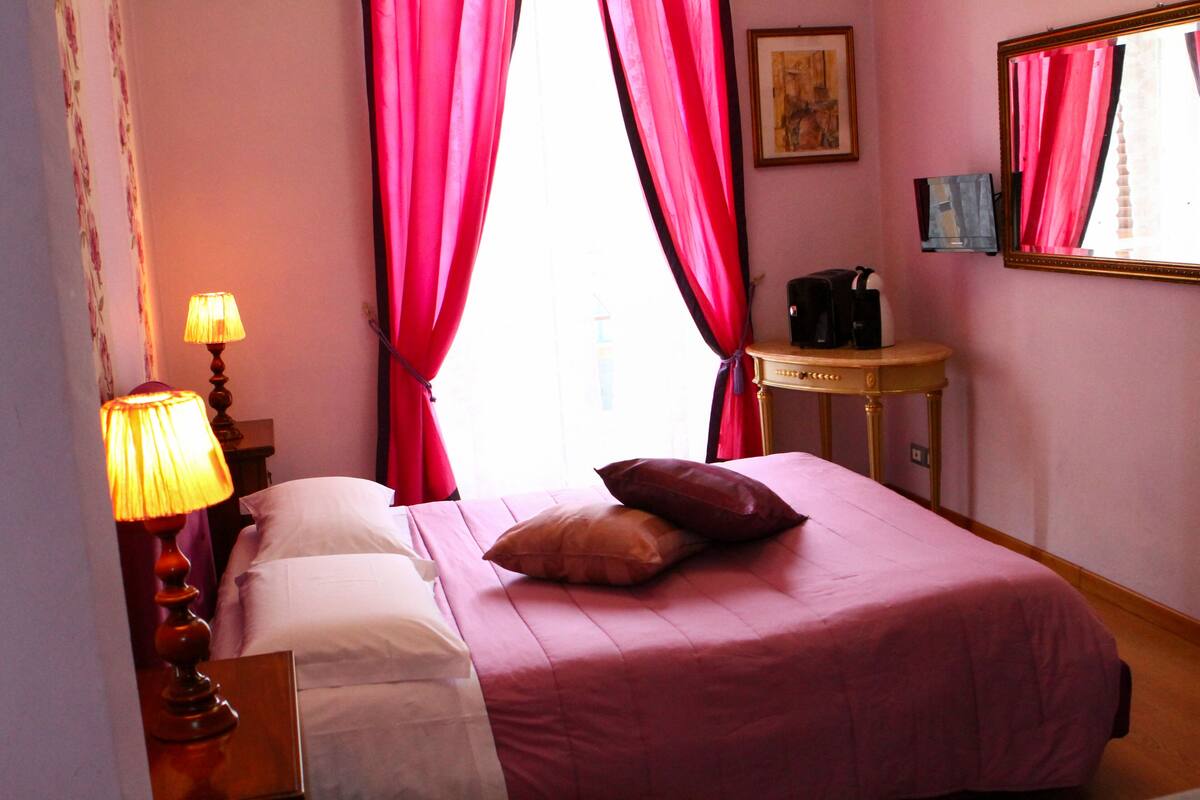 A hotel room with a bed, a nightstand with a lamp, a small table with a coffee maker, and a mirror on the wall.