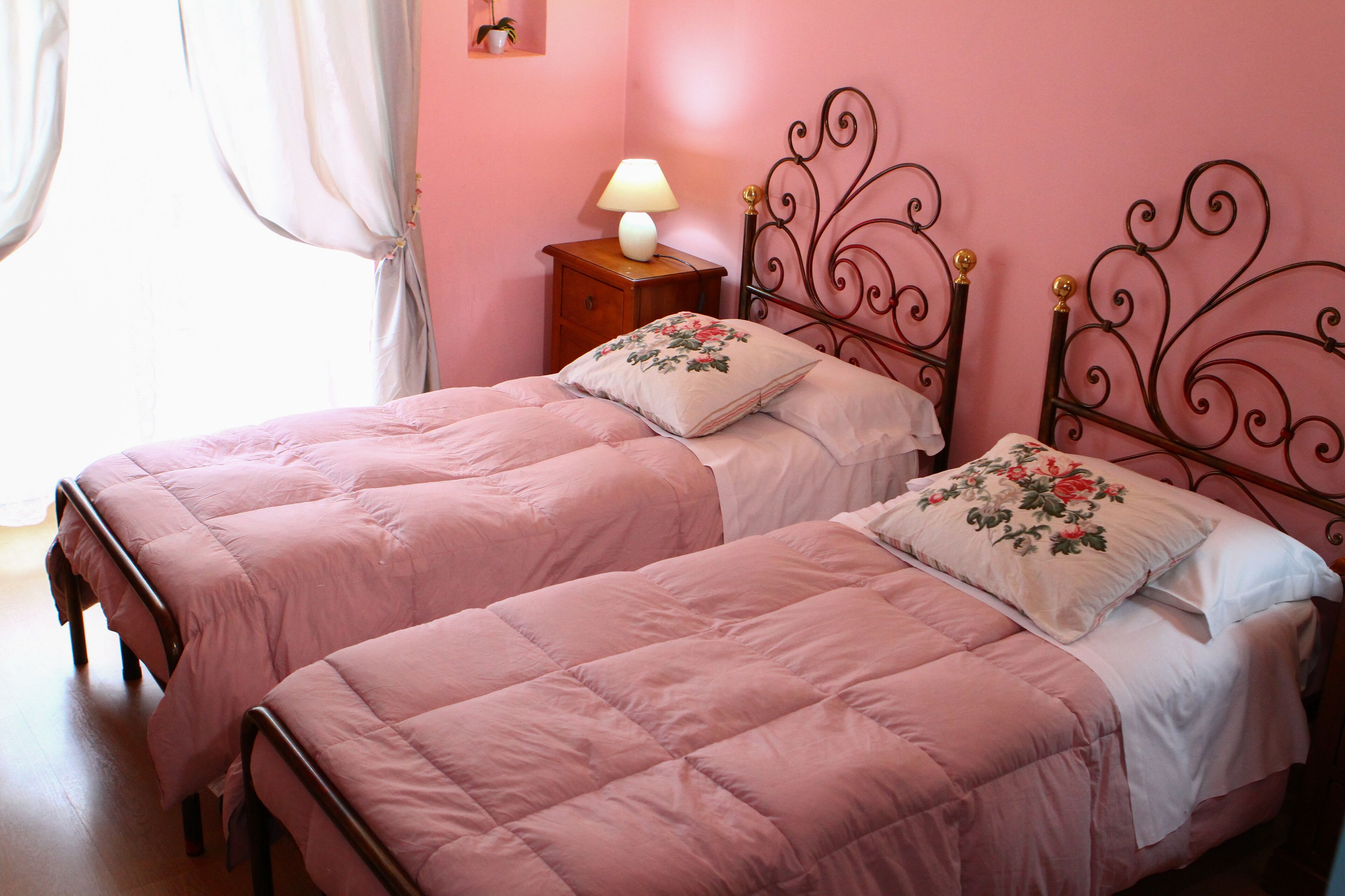 Quarto com duas camas, cada uma com colcha rosa e almofadas florais, uma mesa de cabeceira com abajur, e uma parede rosa.