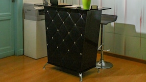 Un mostrador de recepción con una silla negra con capitoné, un jarrón verde con una flor y una computadora portátil. En la pared hay diplomas enmarcados y una puerta azul a la izquierda.