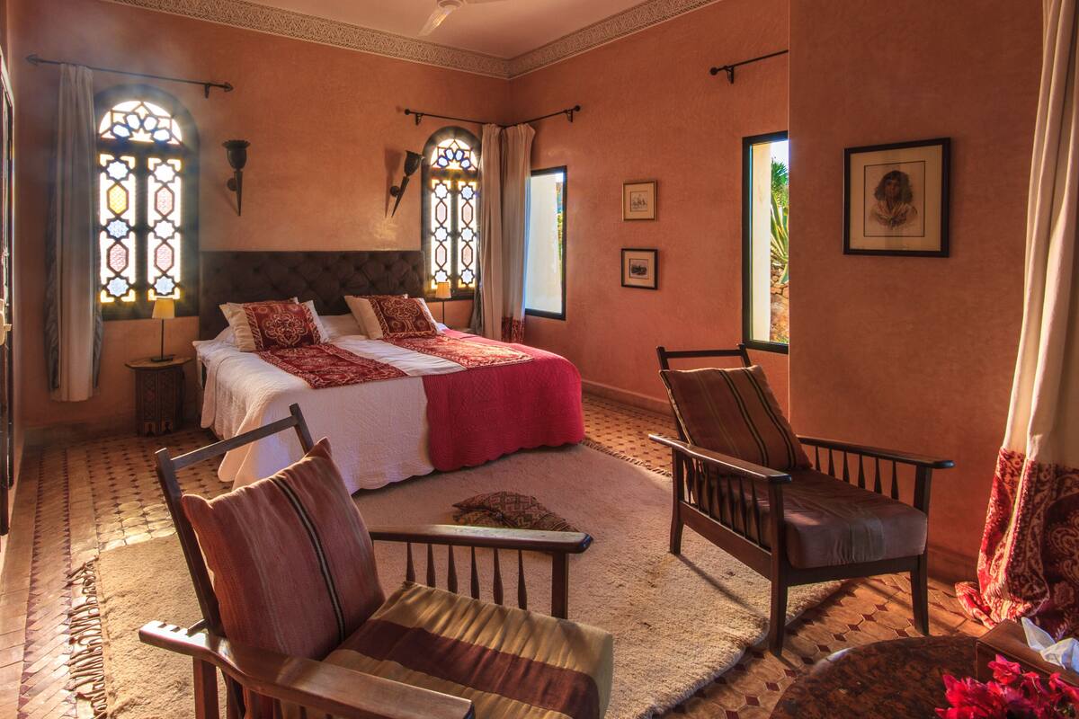 A bedroom with a large bed, two chairs, a nightstand, and a window with curtains.