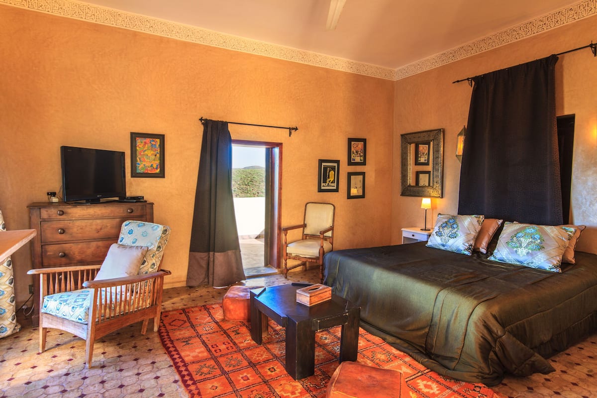A hotel room with a bed, a wooden chair, a small table, a television, and a window with a view of the outdoors.