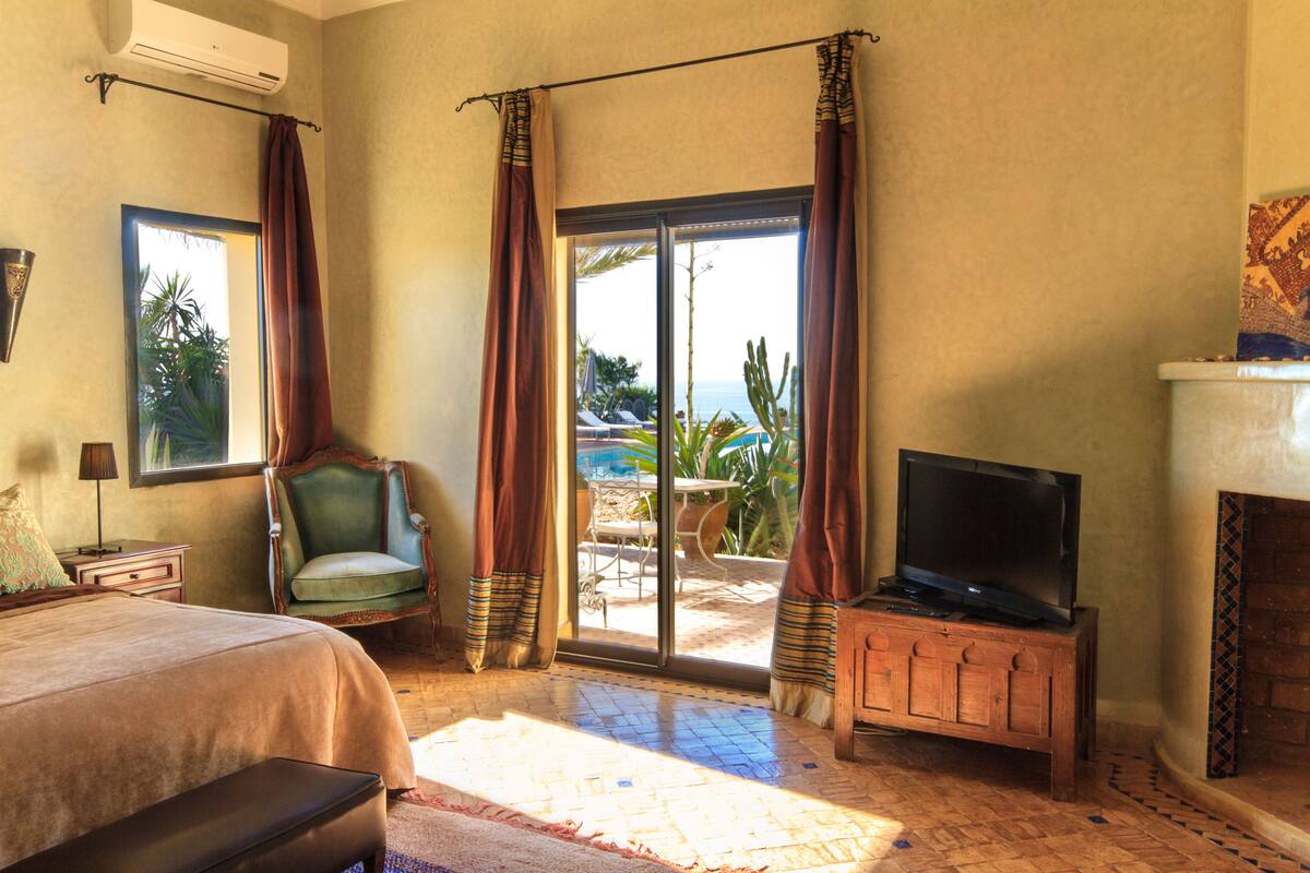 A bedroom with a bed, a television on a wooden stand, a chair, and a view of the outdoors through a sliding door.