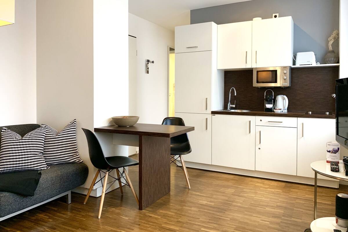 Een ruimtelijke woonruimte met een kitchenette, een eethoek met tafel en stoelen, en een bank met gestreepte kussens.