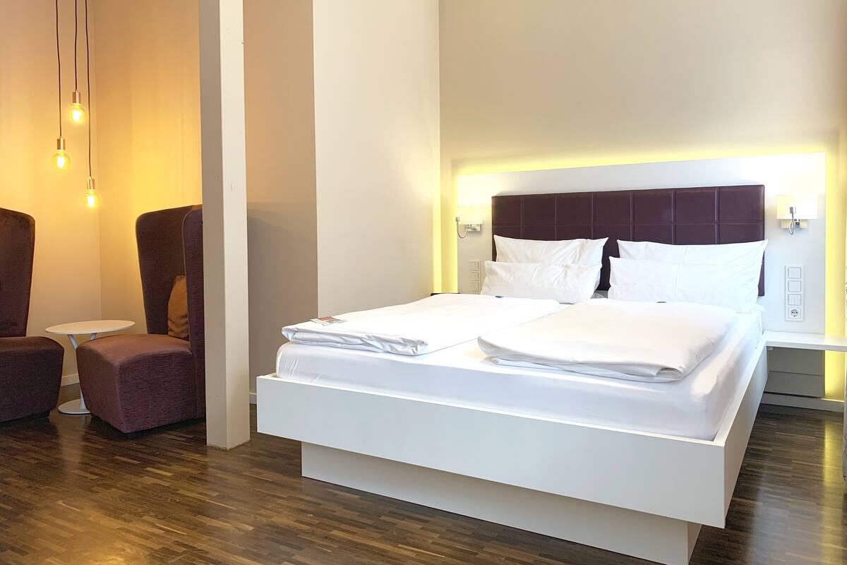 Een moderne hotelkamer met een groot bed, een klein rond tafeltje, twee fauteuils en een houten vloer.