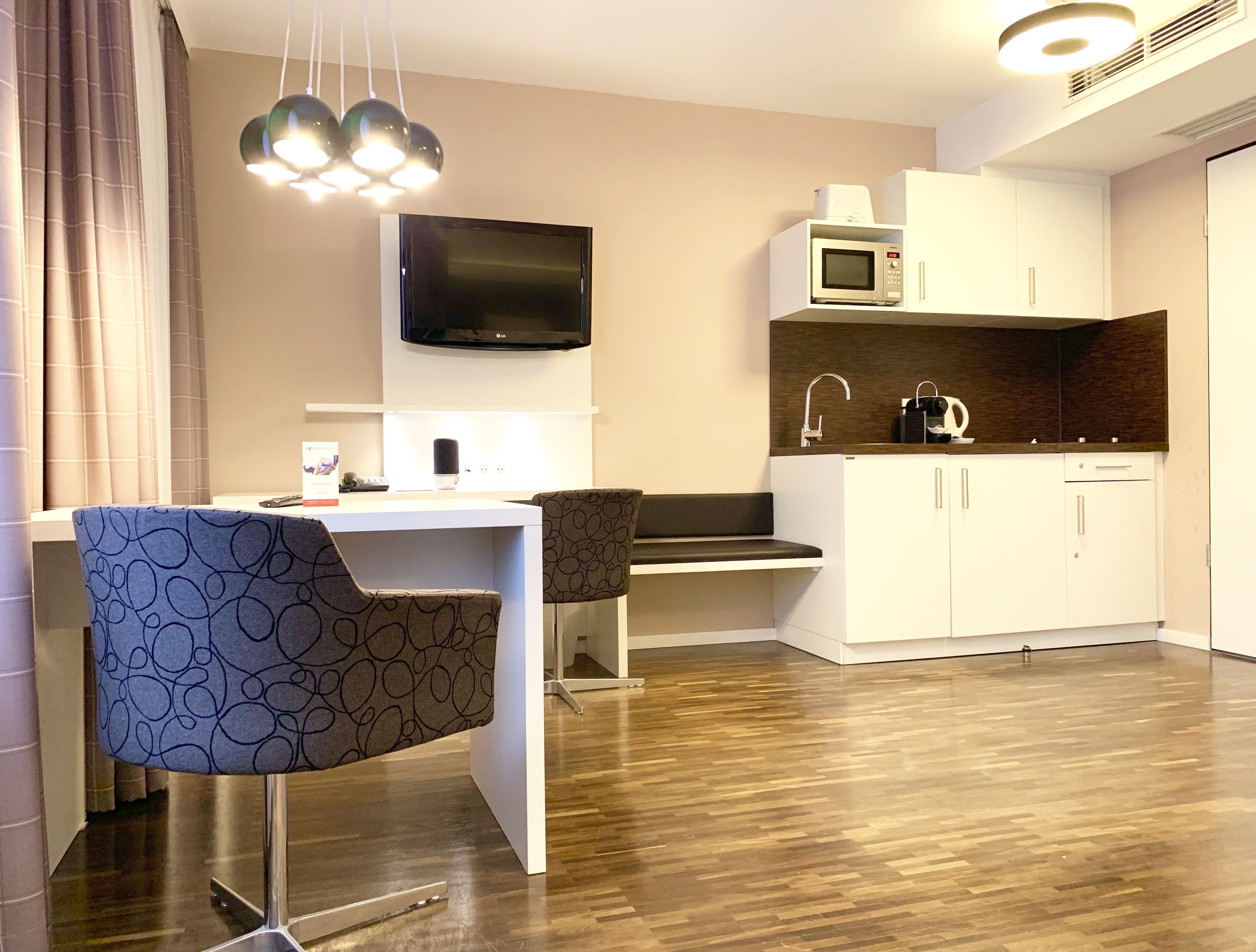 A modern kitchen with a flat-screen TV, microwave, and sink. There is a dining area with a chair and a table.