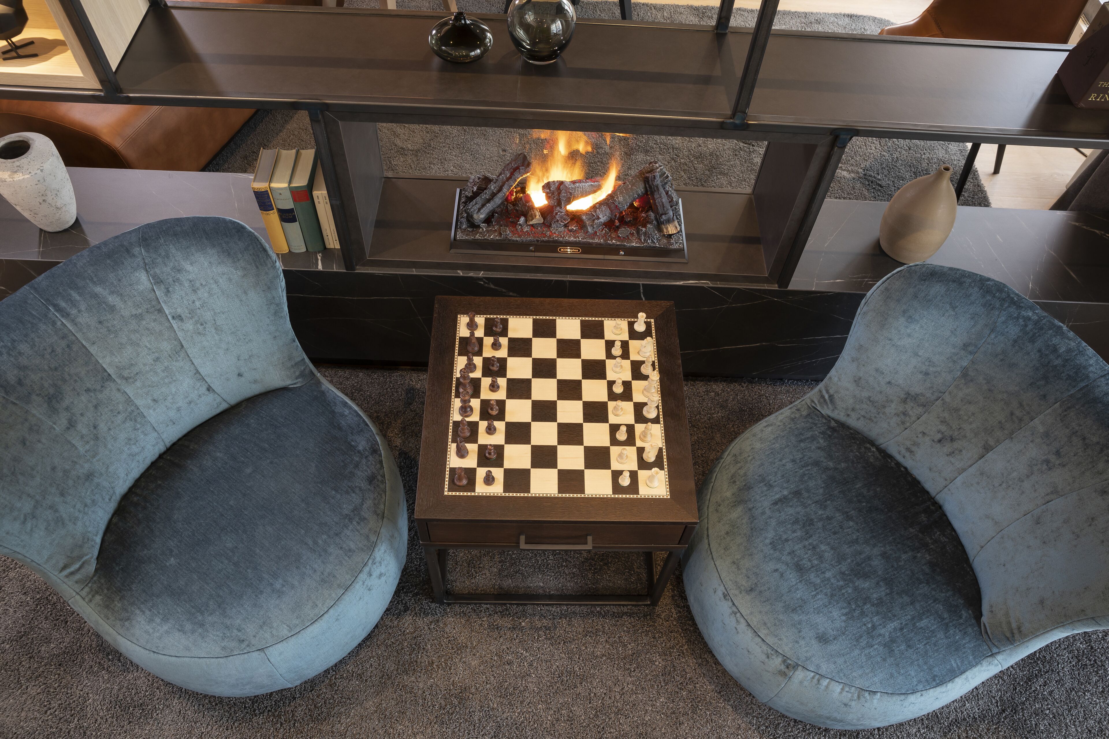 A cozy seating area with two armchairs, a small table with a chessboard, and a fireplace with a fire.
