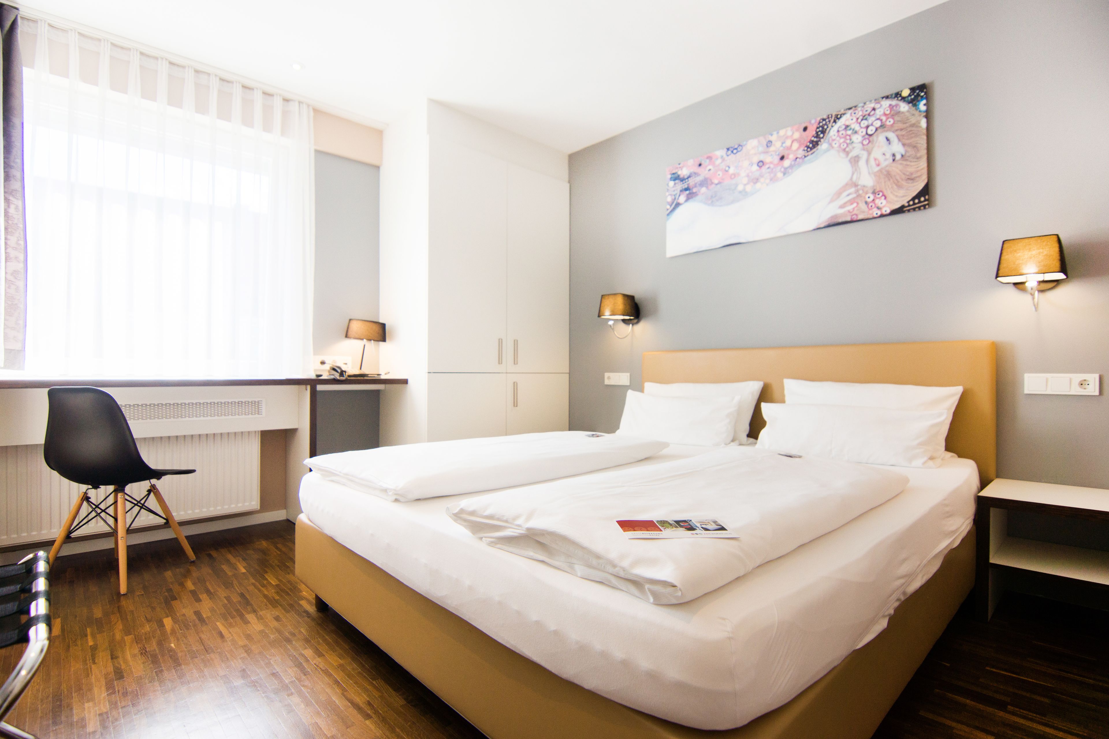 A hotel room with a bed, a desk, a chair, and a painting on the wall.