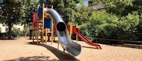 Un parque infantil con tobogán y estructuras para escalar, rodeado de árboles y suelo de arena.