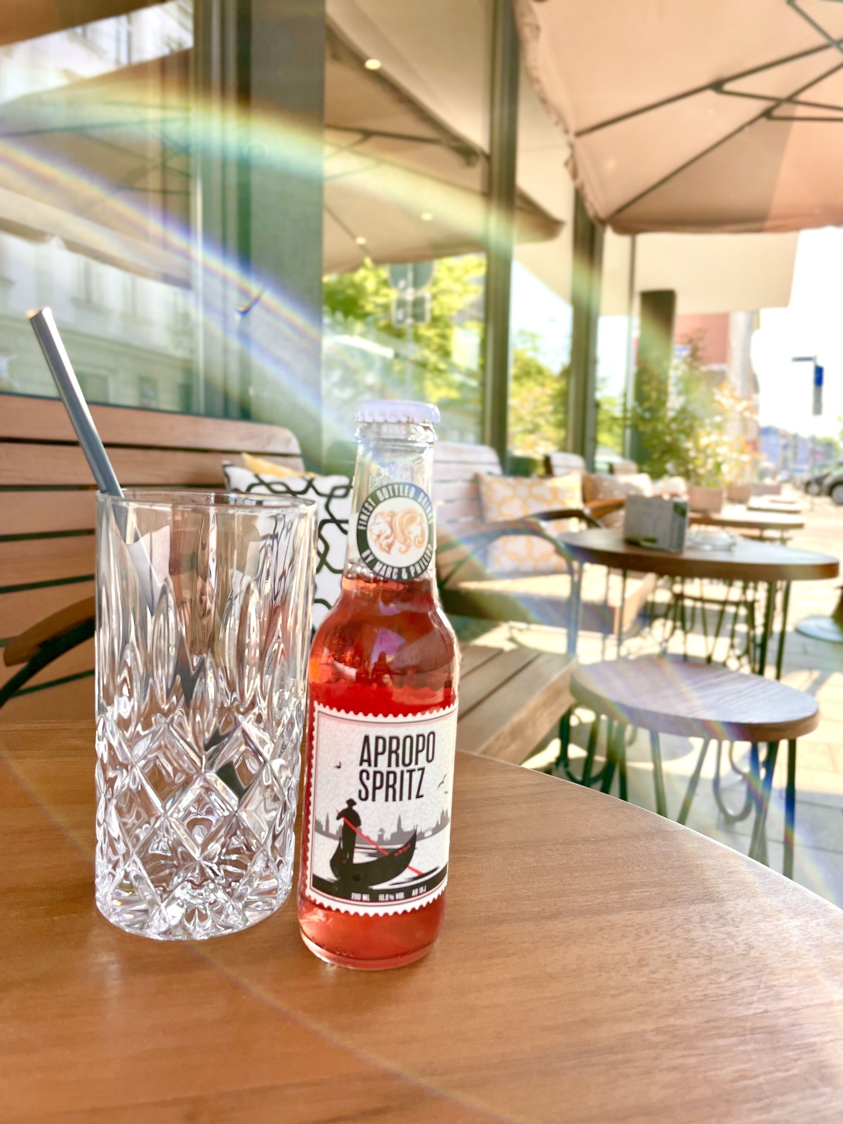 Une bouteille d’Apropo Spritz et un verre avec une paille, posés sur une table en plein air.