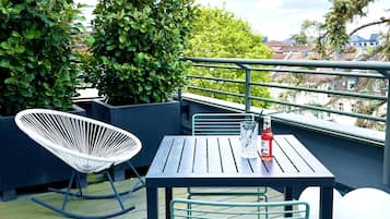 A rooftop terrace with a table and chairs, surrounded by greenery and overlooking a residential area.