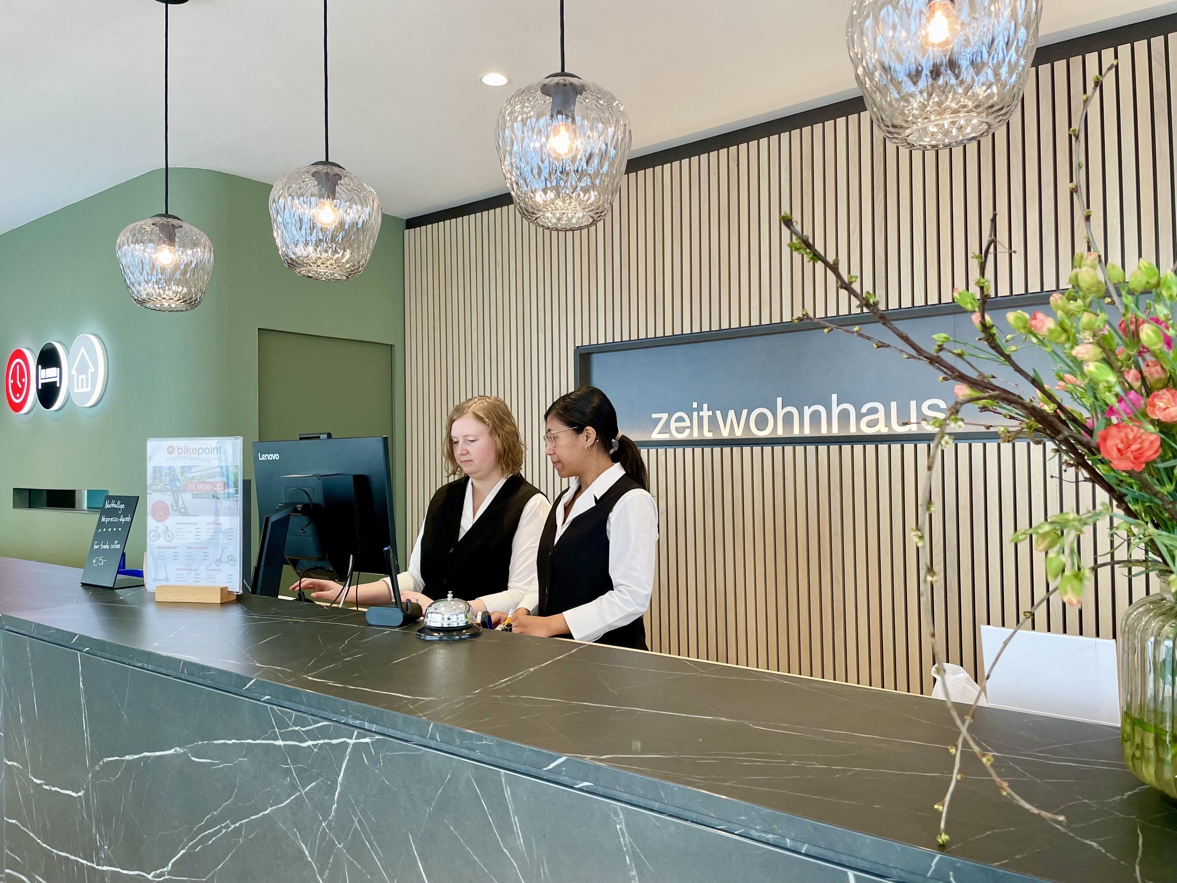 Un comptoir d’accueil au design moderne, doté d’un dessus en marbre et d’un mur revêtu de panneaux en bois. Trois lampes suspendues illuminent l’espace au-dessus du comptoir, et une enseigne portant l’inscription « zeitwohnhaus » est visible.