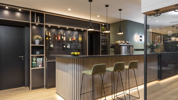 Zona de bar moderna con mostrador de madera, taburetes y una variedad de botellas y vasos.