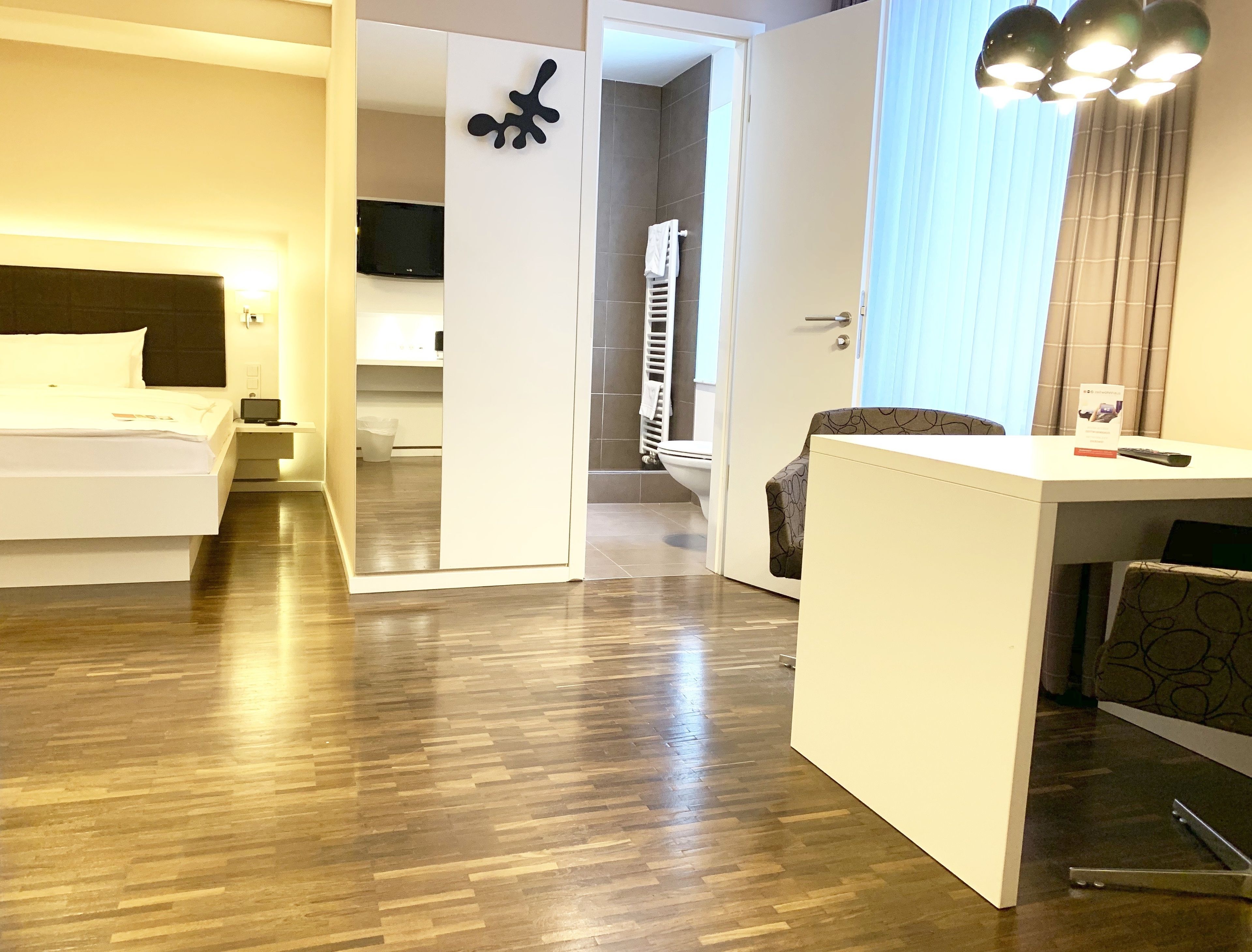 Habitación de hotel con cama, escritorio, silla, televisión y baño con tendedero para toallas.