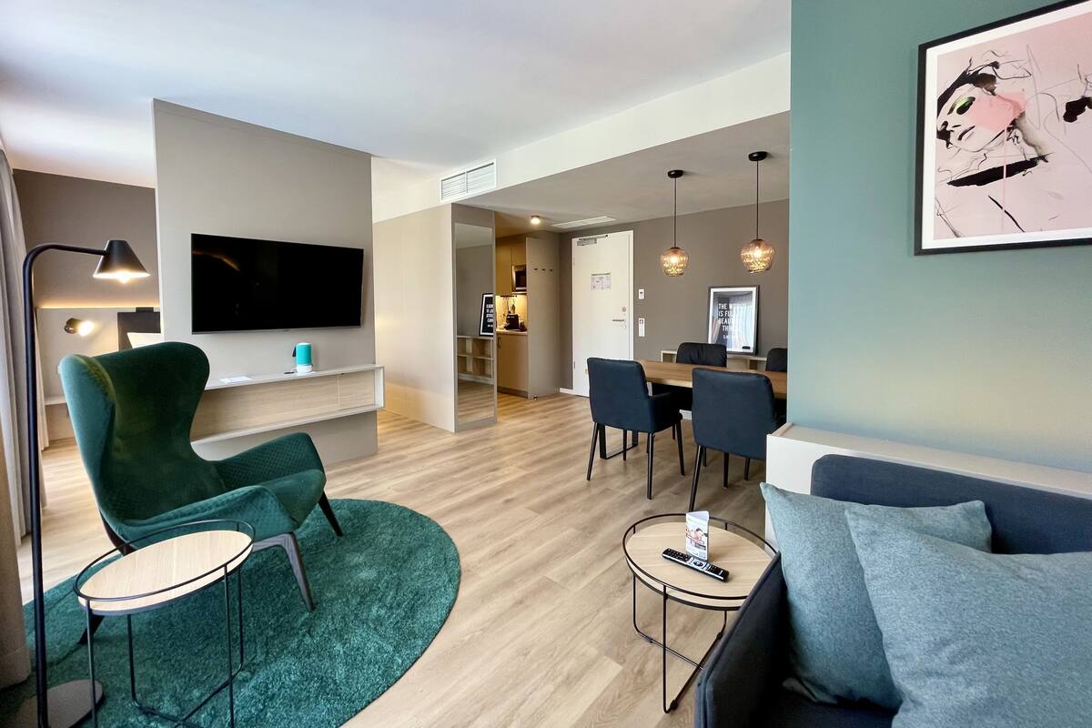 Een moderne woonkamer met een groene fauteuil, een ronde salontafel en een bank. Er is een eethoek met een tafel en stoelen, en een keuken op de achtergrond.