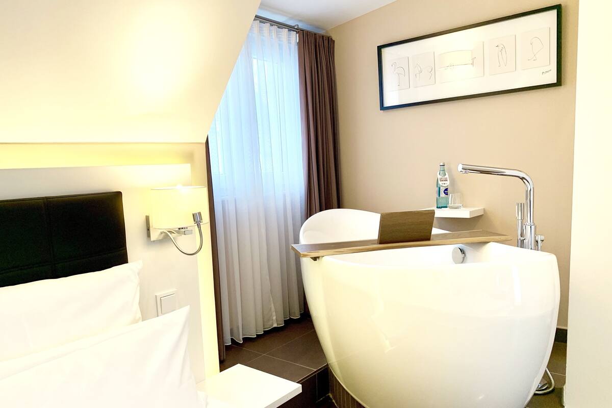 Een moderne badkamer met een groot bad, een douche en een toilet. Er is een bed met witte lakens en een hoofdbord, en een ingelijst kunstwerk aan de muur.