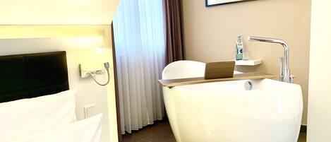 A modern bathroom with a large bathtub, a shower, and a toilet. There is a bed with white linens and a headboard, and a framed picture on the wall.