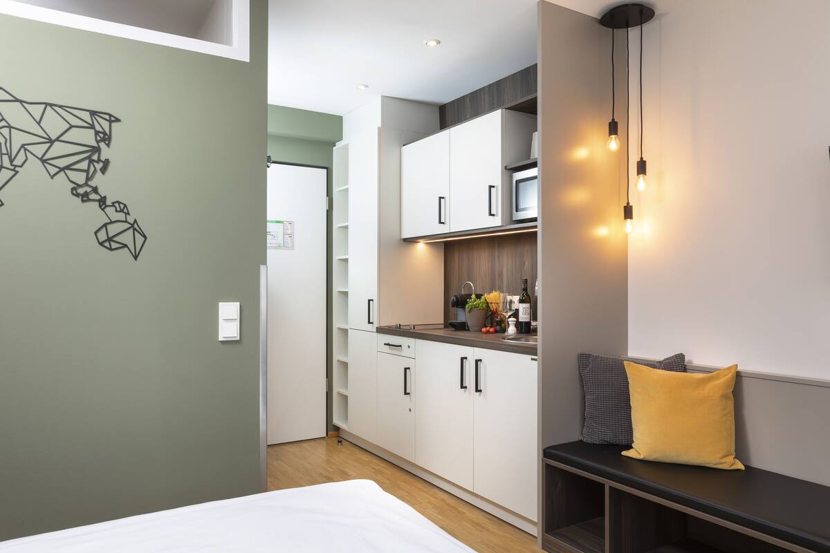 Een moderne keuken met witte kasten, een magnetron en een spoelbak. Er staat een bankje met een geel kussen en een grijs kussen.