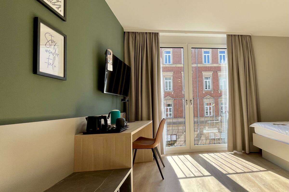 Een moderne hotelkamer met een bureau, een flatscreen televisie, een bed en een groot raam met uitzicht op de stad.