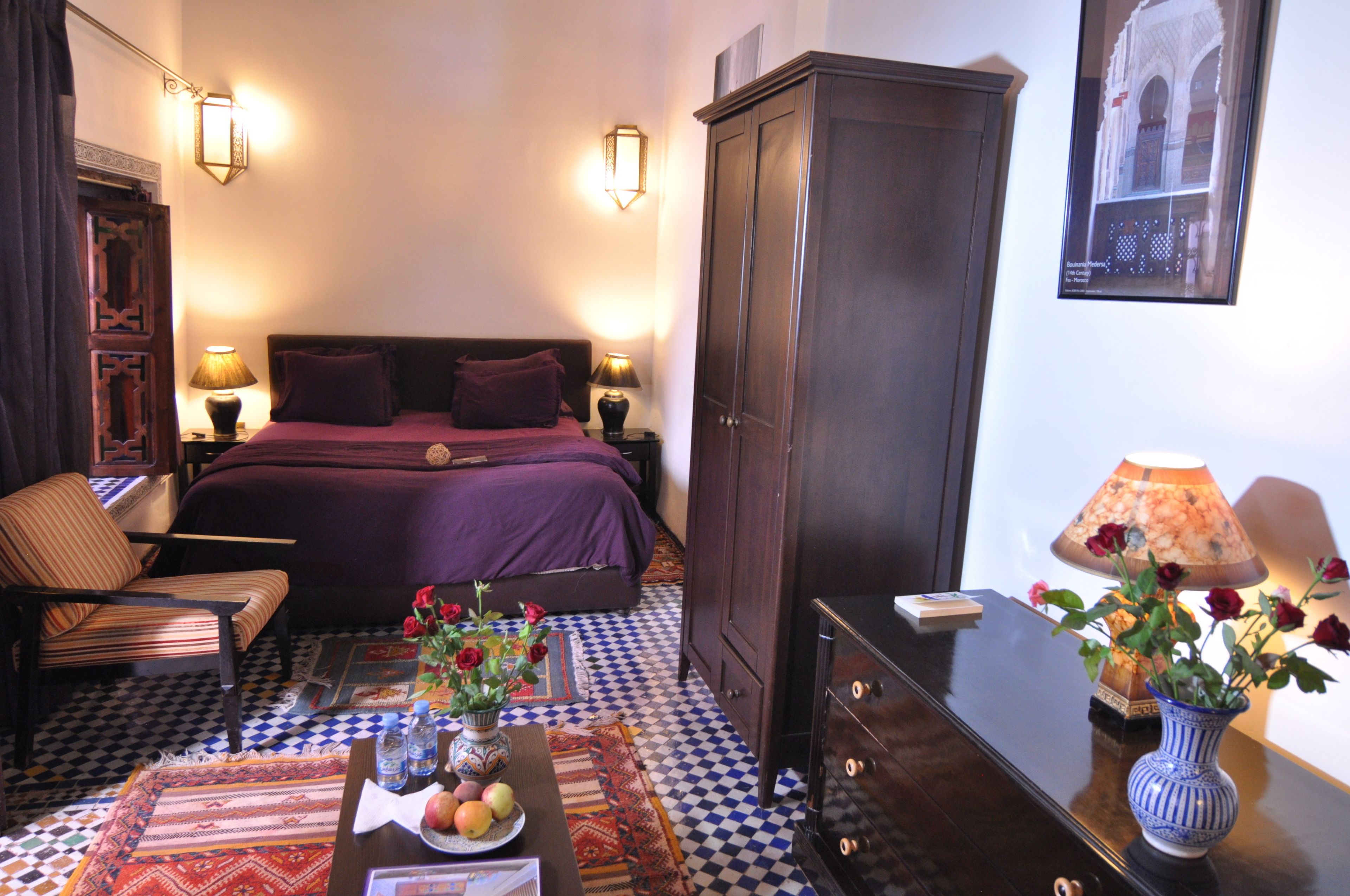 A hotel room with a checkered floor, a bed with purple bedding, a wooden wardrobe, a chair, a table with a vase of flowers, and wall-mounted lamps.