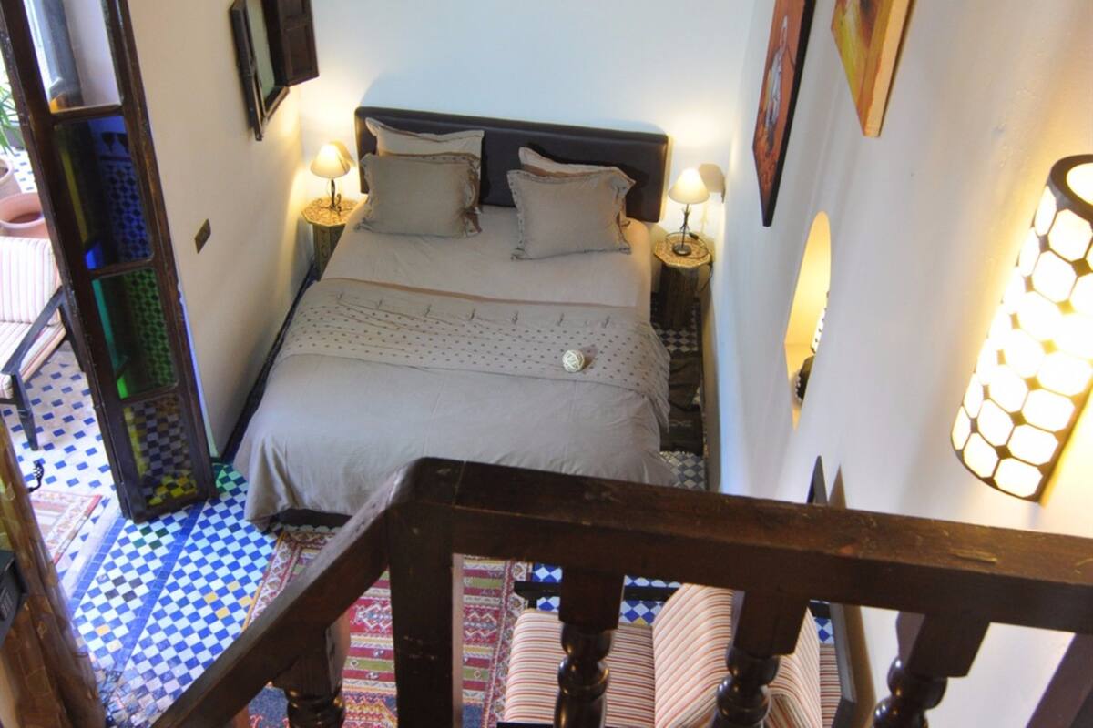 Una escalera estrecha conduce a una cama con suelo a cuadros, cabecero de madera y dos lámparas de noche.