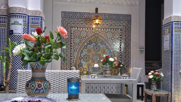 A room with a patterned tile wall, a table with a vase of flowers, and a chair. - Riad Adarissa (Fes)