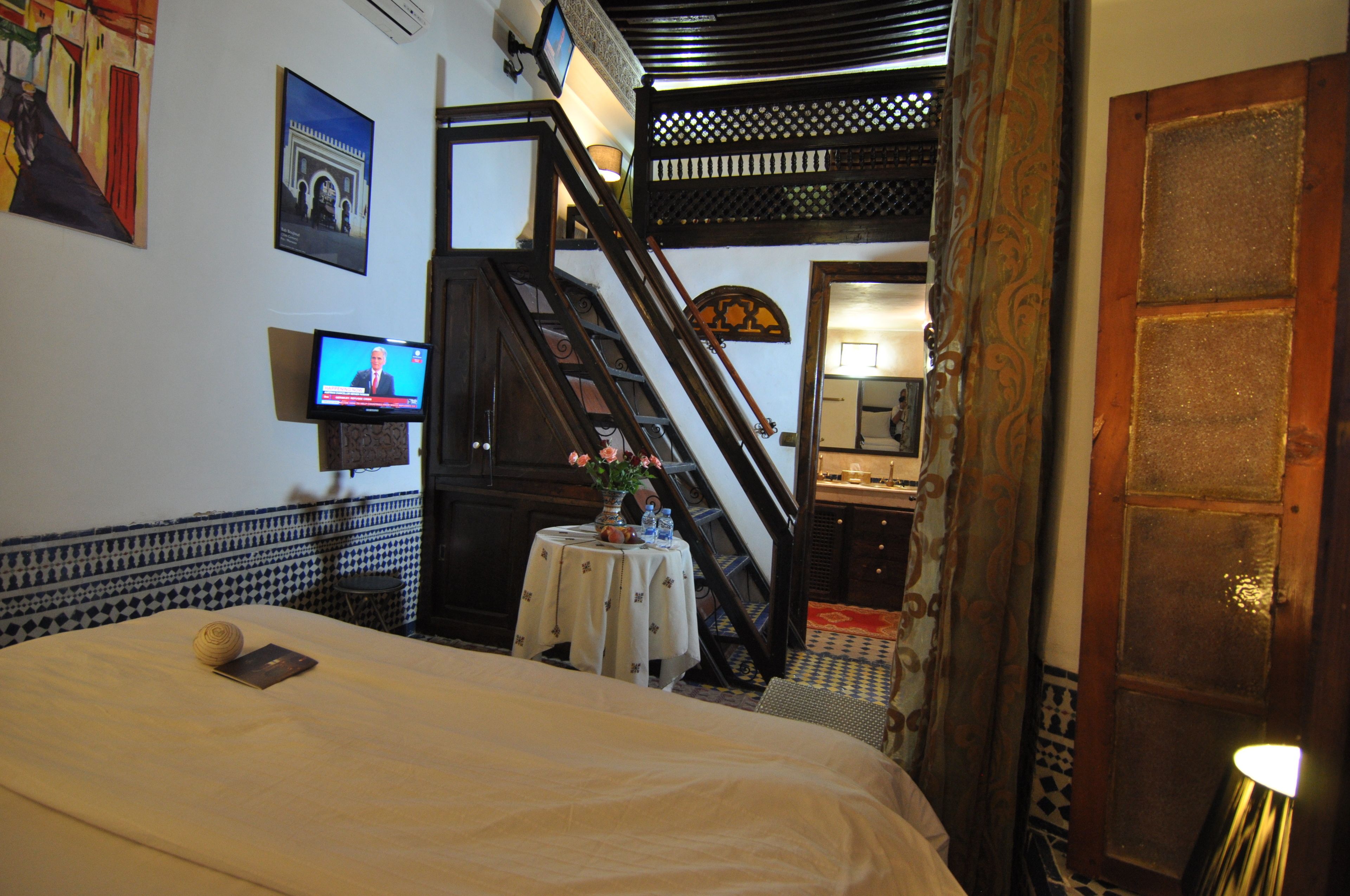 A hotel room with a bed, a television, a small table with a vase, a staircase, and a door.