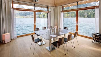 Un salón de conferencias con una mesa, sillas y un proyector. Cuenta con ventanas amplias con cortinas, piso de madera y vistas a un lago y montañas.
