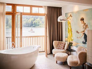 A bathroom with a freestanding bathtub, a chair, a table with a vase, and a painting on the wall.