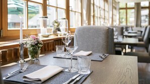 A dining table set with cutlery, glasses, and a candle, in a room with large windows and a bench in the background.