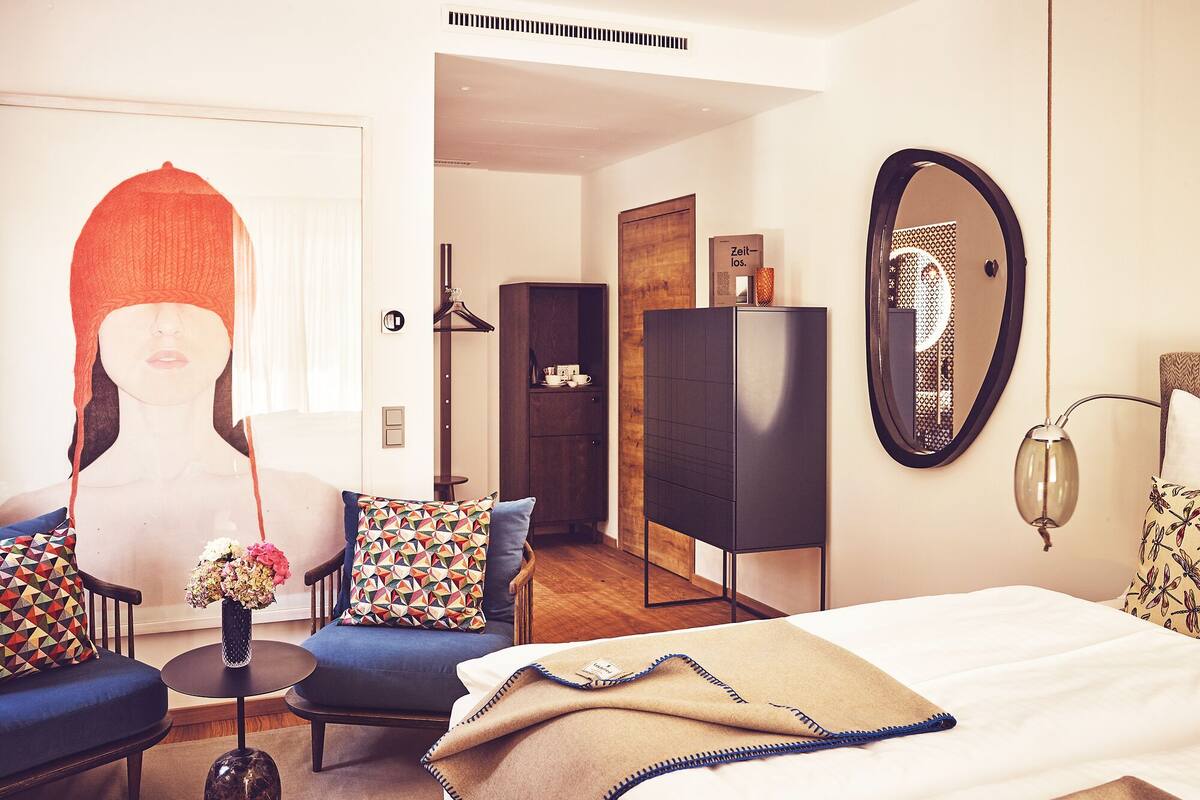 Ein modernes Hotelzimmer mit Bett, einer Couch, einem Sessel, einem kleinen Tisch und einem Spiegel.