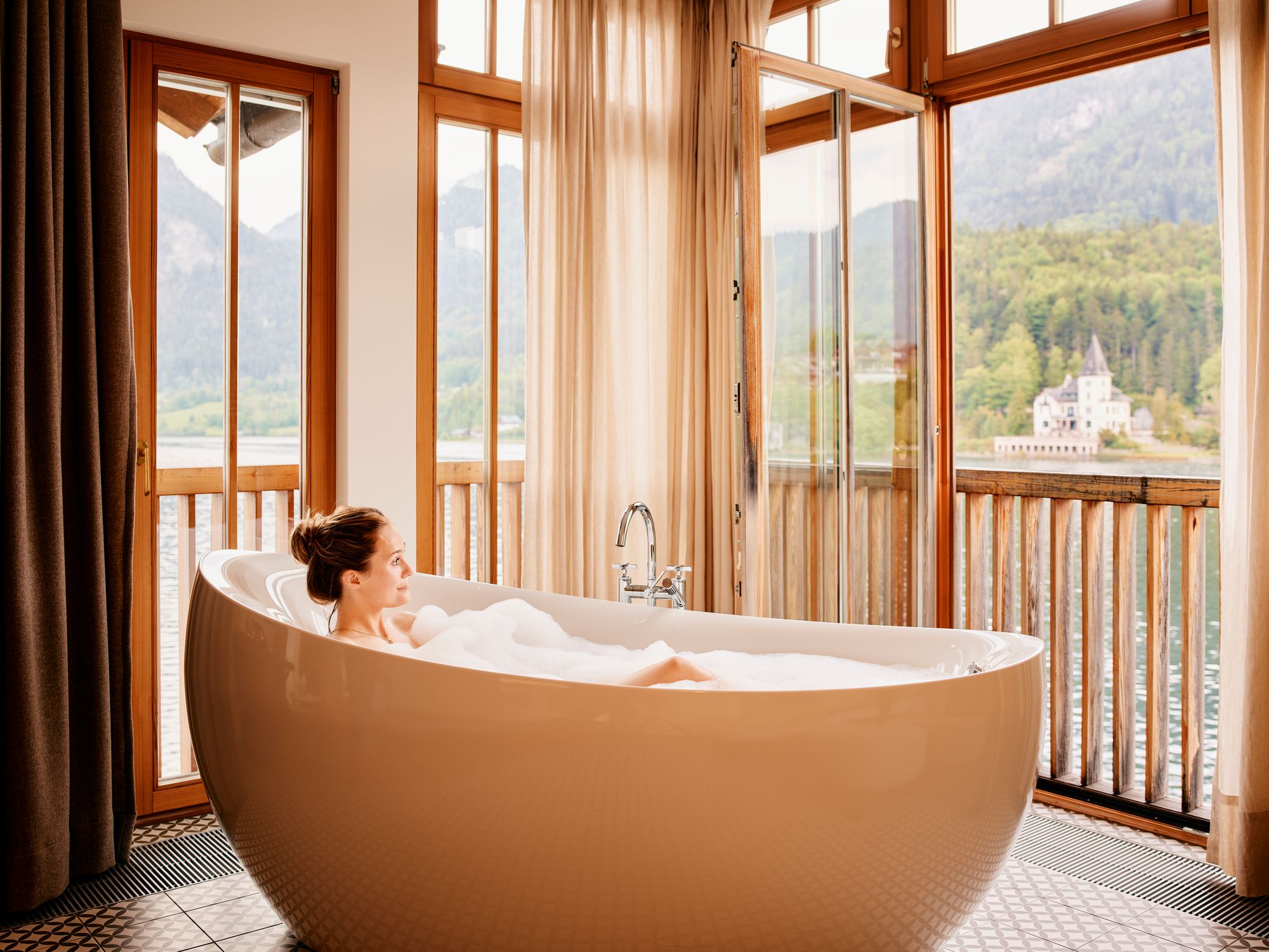 A woman relaxing in a large bathtub with a view of a castle through the windows.
