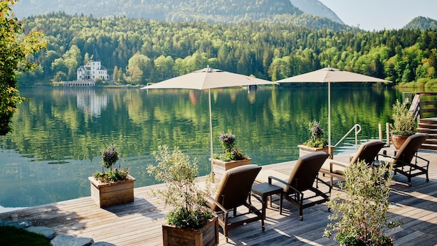 Terraza de madera con mobiliario exterior y sombrillas, con vistas a un lago y una casa al fondo.