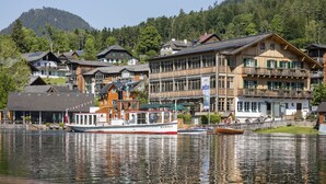Un barco de madera llamado "RUDOLF" amarrado en un lago frente a un hotel con edificios de varios pisos y una montaña boscosa al fondo.