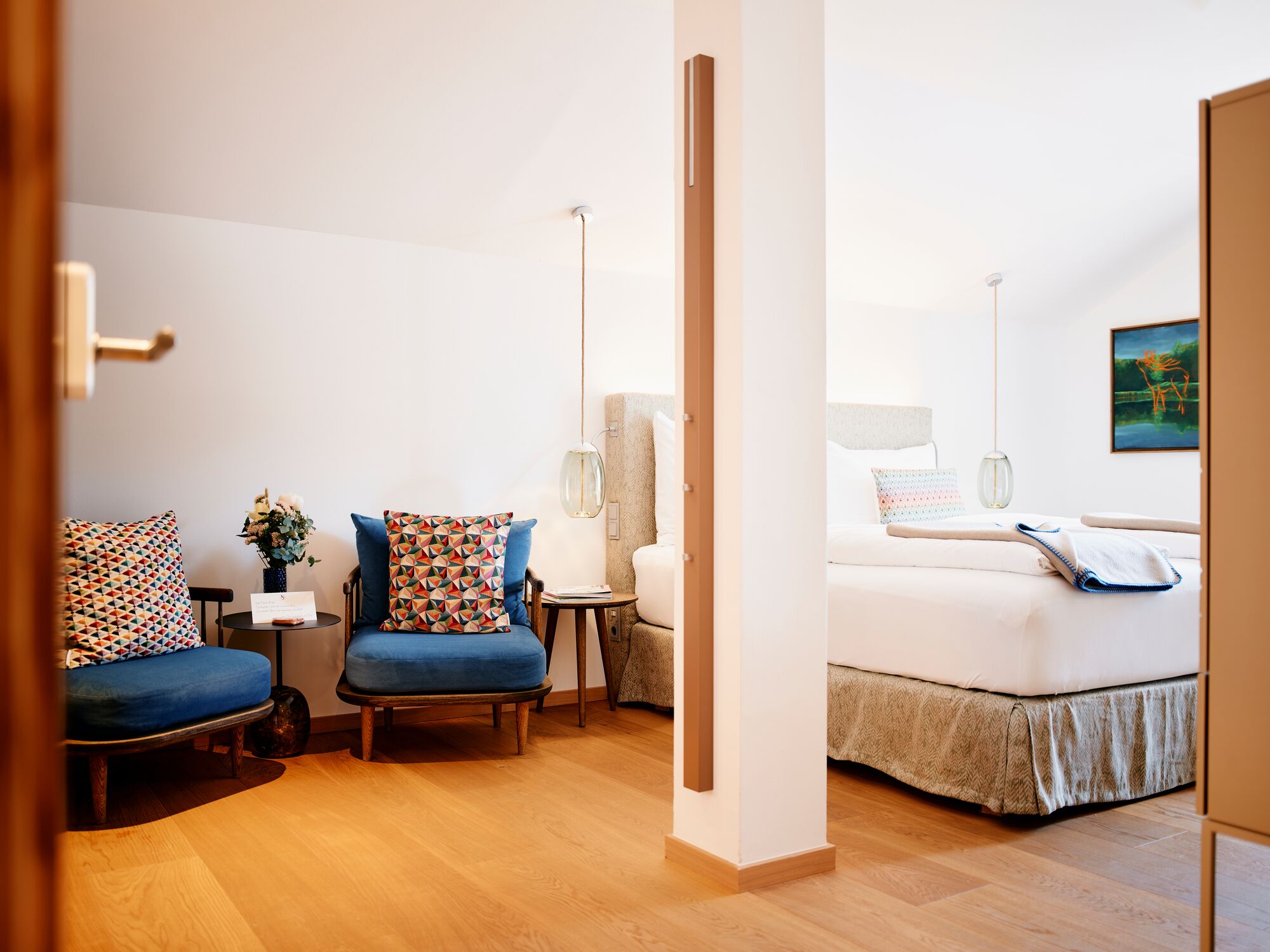A hotel room with a bed, two blue armchairs, a small table with a vase, and a lamp.