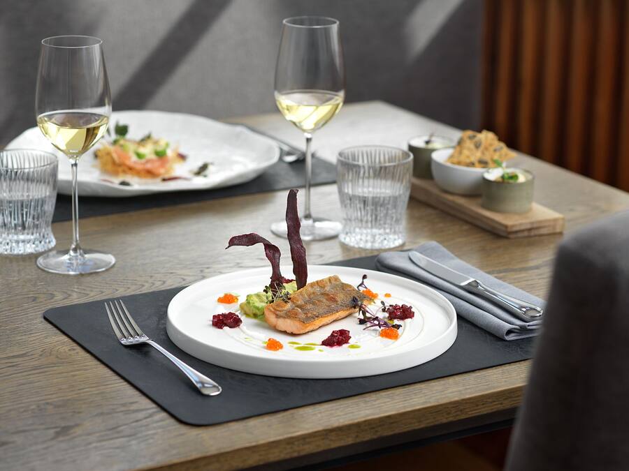 A dining table with a white plate of food, two glasses of white wine, and a napkin with cutlery.