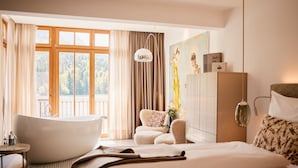 Un dormitorio con una bañera grande, una cama, un sofá y vistas a un bosque a través de amplios ventanales.