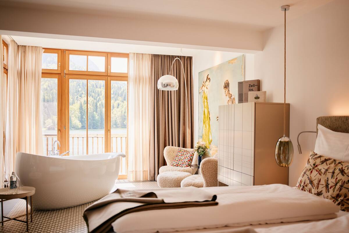 Ein Schlafzimmer mit einer großen Badewanne, einem Bett, einer Couch und Blick auf einen Wald durch große Fenster.