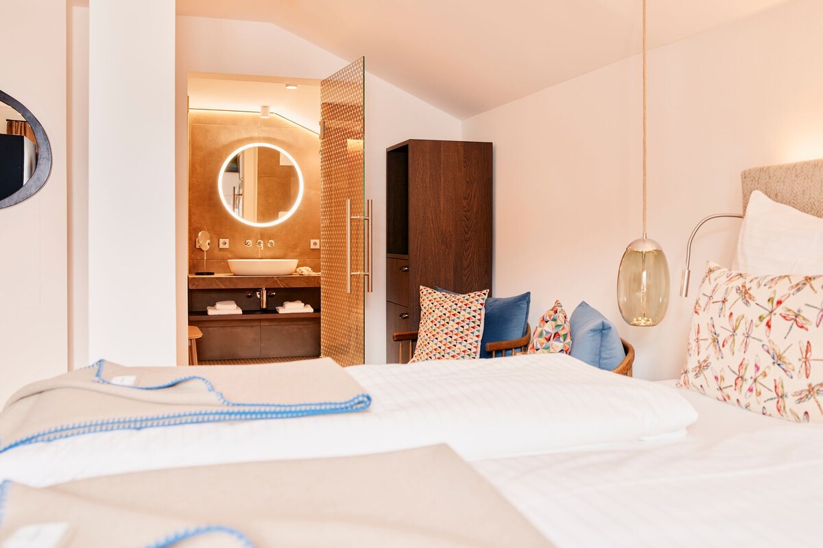 Habitación de hotel con cama, mesita de noche y baño con espejo, lavabo y toallero.