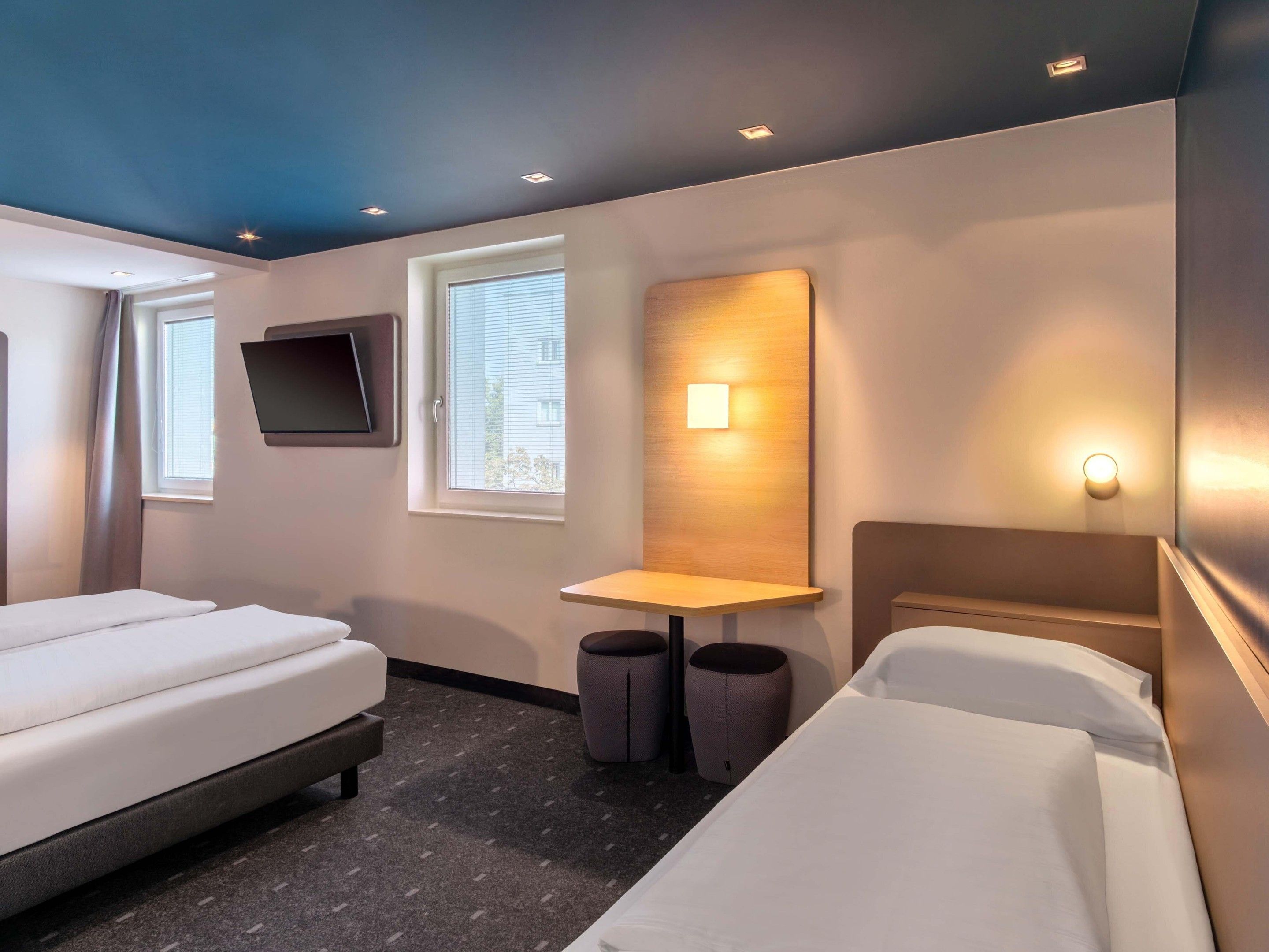 Habitación de hotel con dos camas, un escritorio, una silla, un televisor montado en la pared y una ventana con persianas.