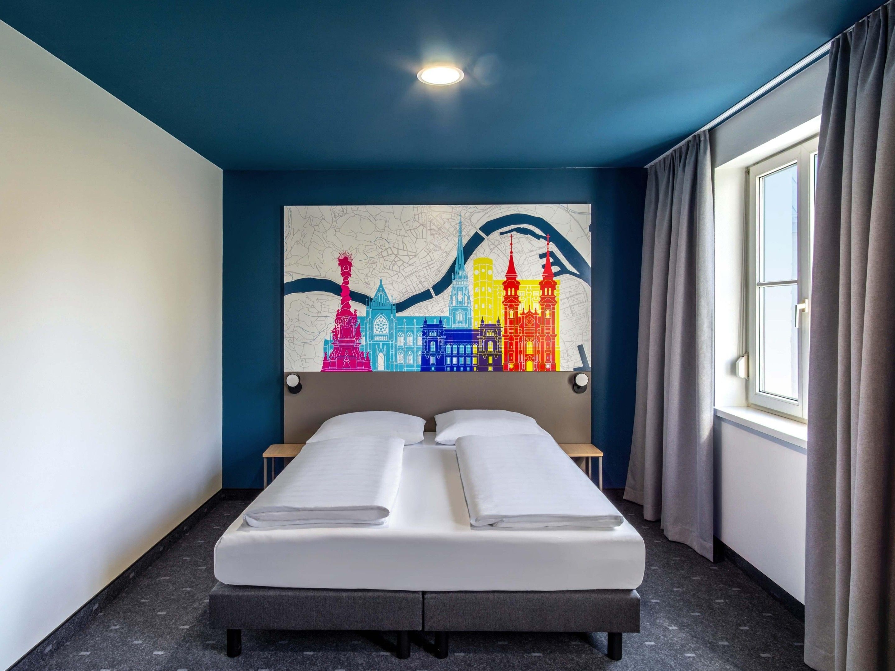 Ein Hotelzimmer mit Bett, Stadtbild-Kunstwerk an der Wand, Fenster mit Vorhängen und einer Karte an der Decke.