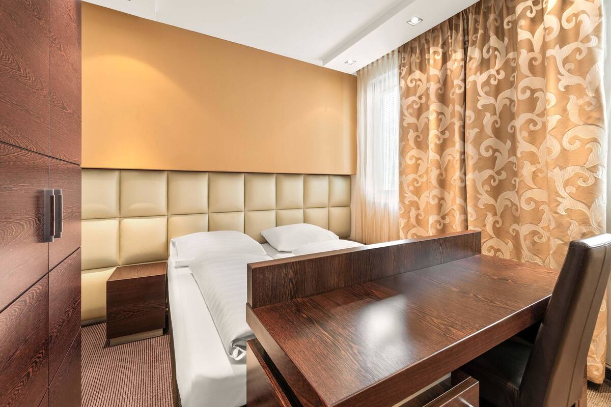 A hotel room with a bed, a wooden desk, a chair, a nightstand, and a large window with curtains.
