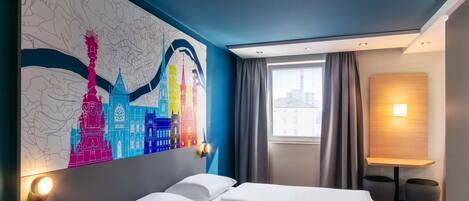 A hotel room with a bed, a nightstand, a wall art of city buildings, a window with curtains, and a bedside lamp.