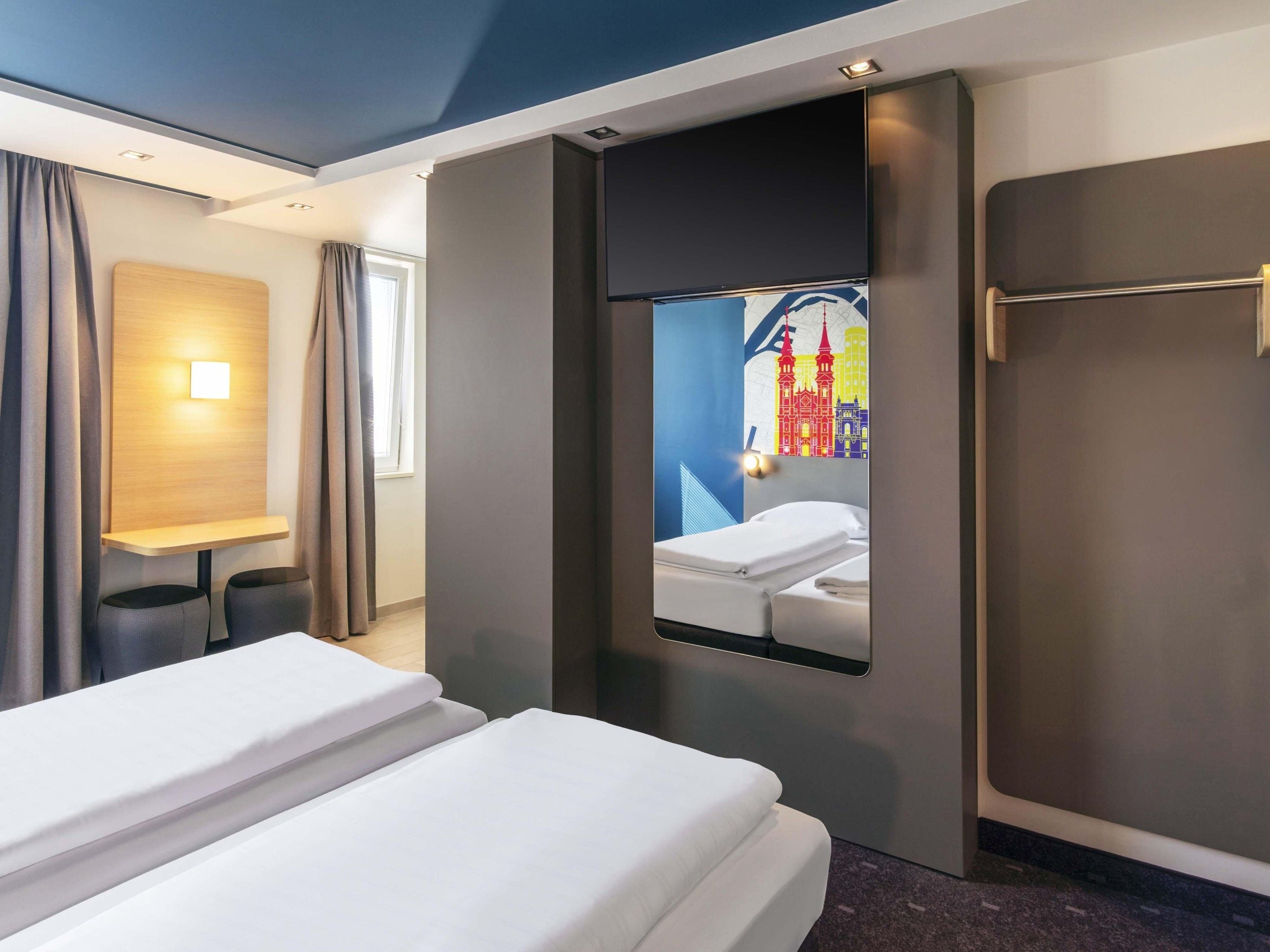 A hotel room with a bed, a desk, and a mirror reflecting a colorful cityscape.