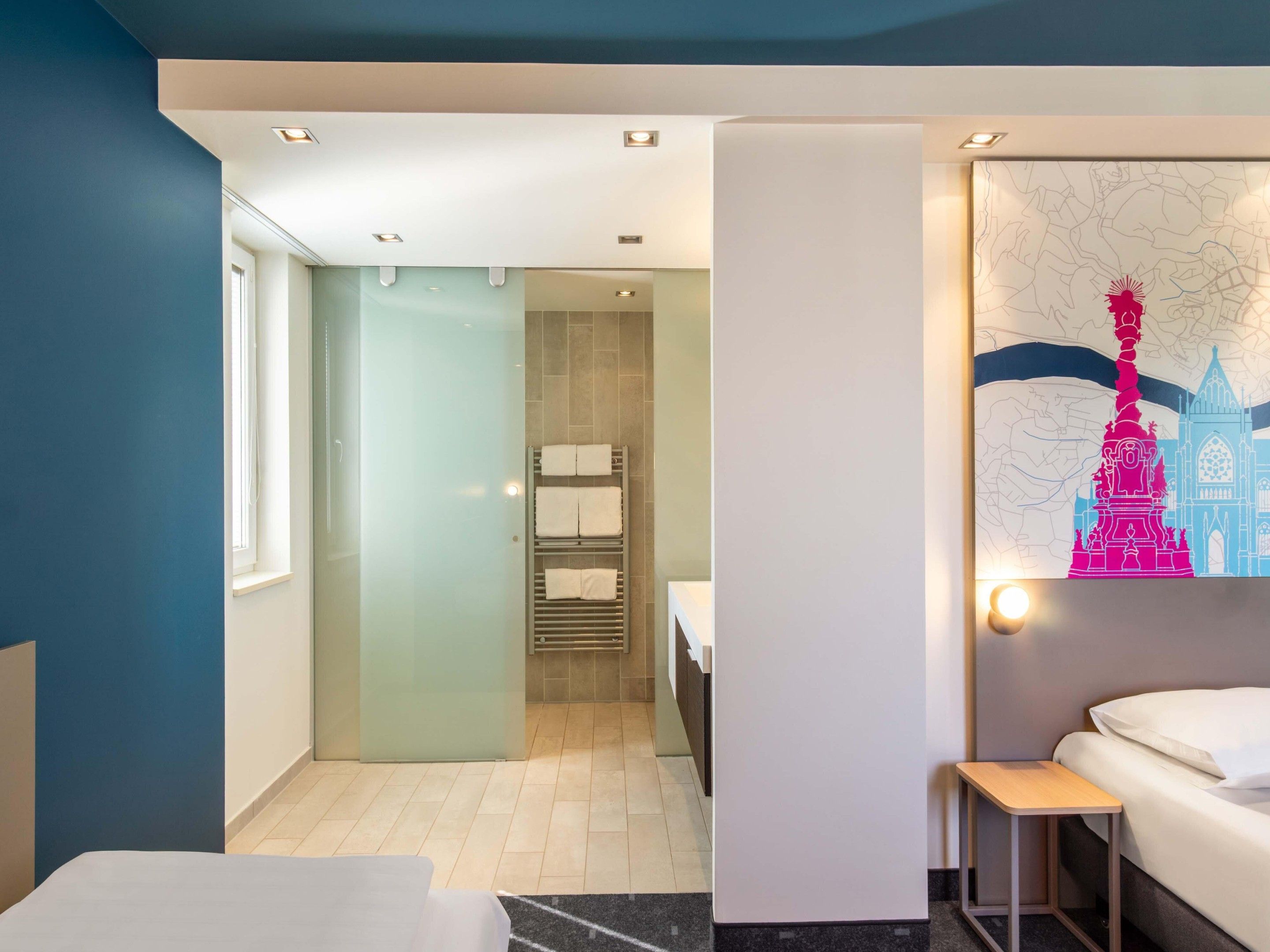 Ein Hotelzimmer mit Bett, einem kleinen Tisch, einem an der Wand hängenden Kunstwerk und einem durchgegangenen Tür erkennbaren Badezimmer.