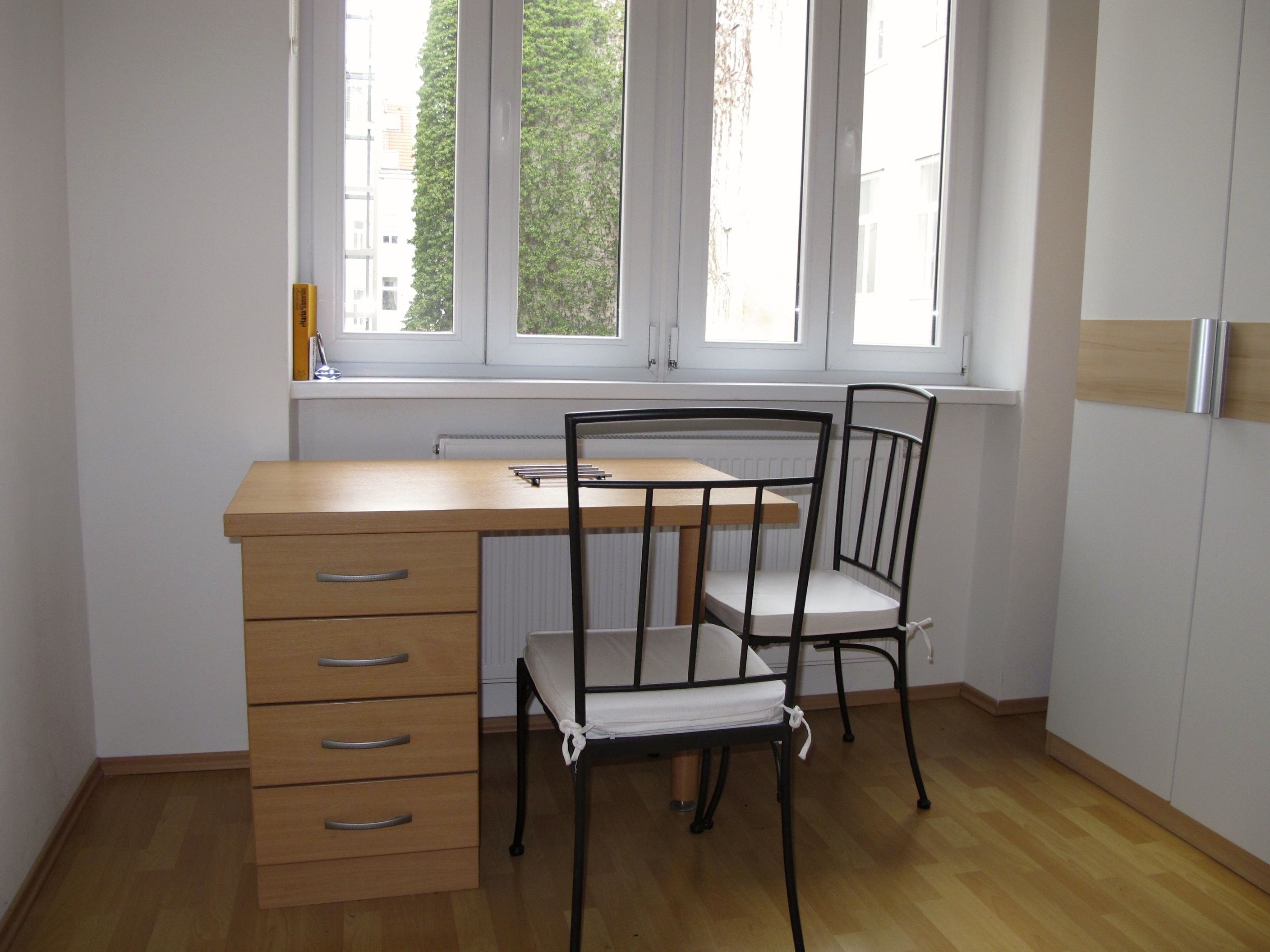 A room with a wooden desk, a chair, a radiator, and a window with a view of buildings.
