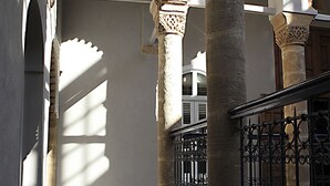 A narrow corridor with stone columns and a tiled floor, leading to a room with a barred window.